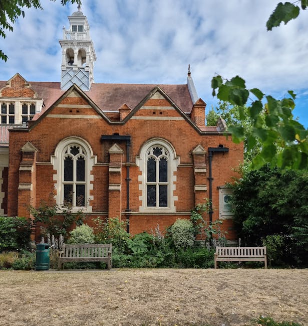 A view of Severndroog Castle garden showing well-maintained flower beds with lush green foliage, including flowering shrubs and small trees, situated in front of a historic red-brick building with dis