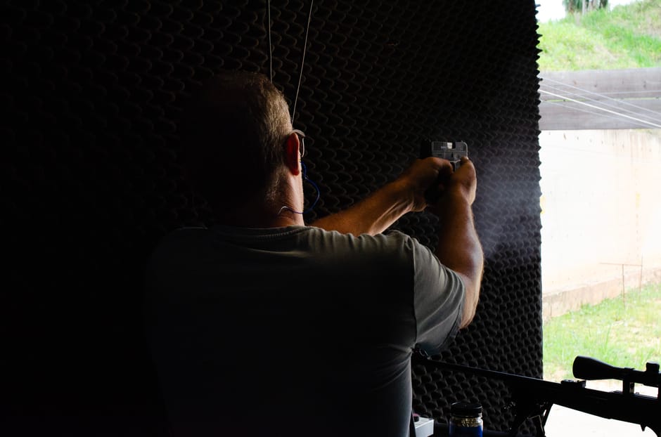 A man with short hair and glasses, dressed in a grey T-shirt, is inside a dark room with soundproof foam panels on the walls, capturing a photo or video with his smartphone. Outside a window, a bright
